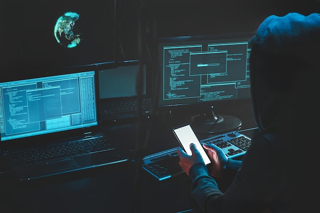 Overhead hacker in hood working at laptop and mobile phone typing text in dark room An anonymous hacker uses malware with mobile phone to hack password personal data steals money from bank cyber