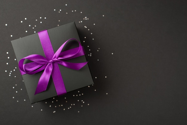 Overhead photo of black giftbox with violet ribbon wrapped as bow confetti and glitter around isolated on the black background