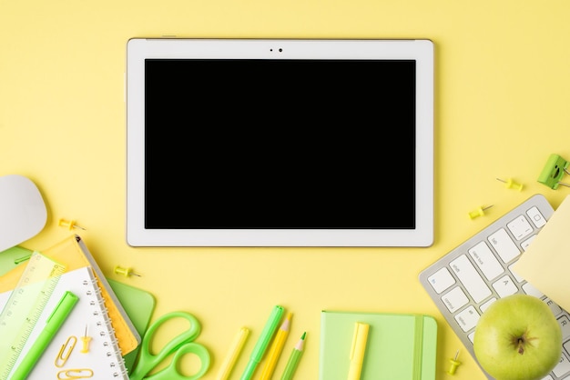 Overhead photo of tablet notebook pen pencil felttip scissors ruler paperclips apple computer mouse and keyboard isolated on the yellow background