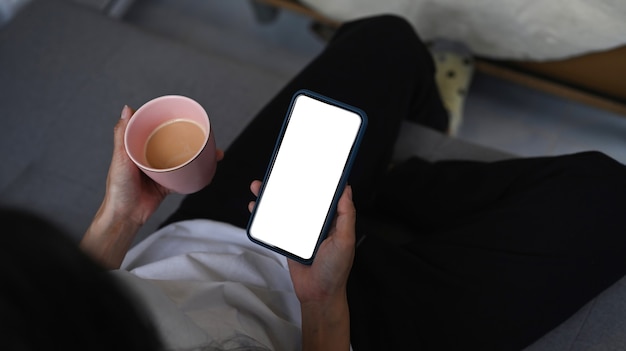 Overhead shot of casual young woman sitting on sofa in living room using smart phone and drinking coffee.