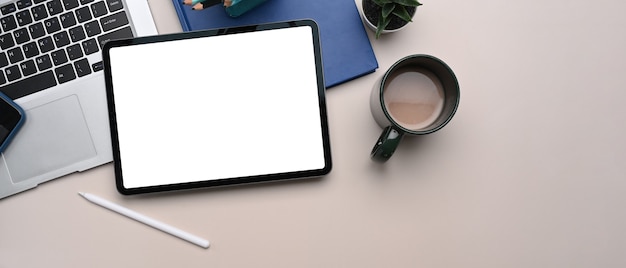 Overhead shot of digital tablet with empty screen, laptop, coffee cup, and notebook on beige cream background.