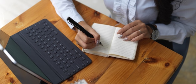 Overhead shot of female office worker writing on blank notebook while working with digital tablet