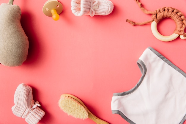 An overhead view of baby's bib; pacifier; sock; brush; stuffed pear and toy on peach background