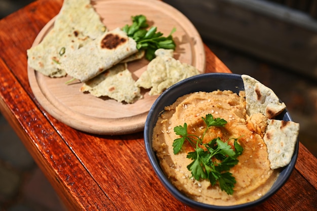 Overhead view of a bowl of chickpea hummus and a wooden board with parsley wrapped in pit bread on a wooden table