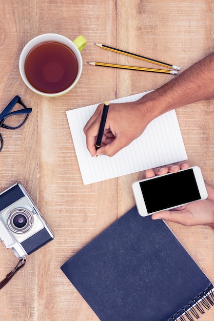 Overhead view of businessman writing on notepad while holding smart phone by camera and coffee on table