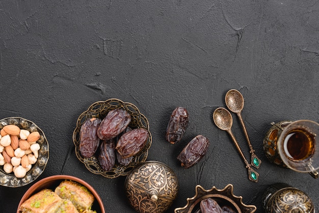 Photo an overhead view of dates in the traditional turkish metallic bowl; spoons and tea glasses on black backdrop