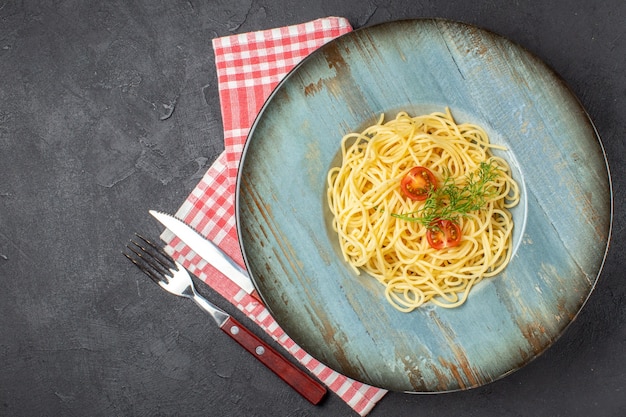 Overhead view of delicious spagetti served with tomatoes greens and cutlery set on red stripped towel on the left side on black background