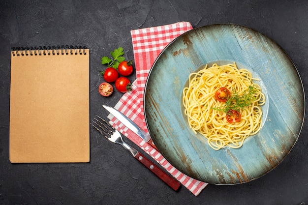 Overhead view of delicious spagetti served with tomatoes greens and cutlery set on red stripped towel notebook on black background