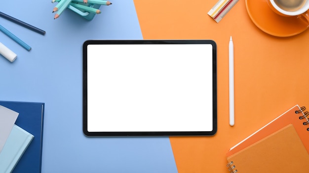 Overhead view of mock up digital tablet with blank screen surrounded by coffee cup, notebook and stationery on two tone blue and orange background.