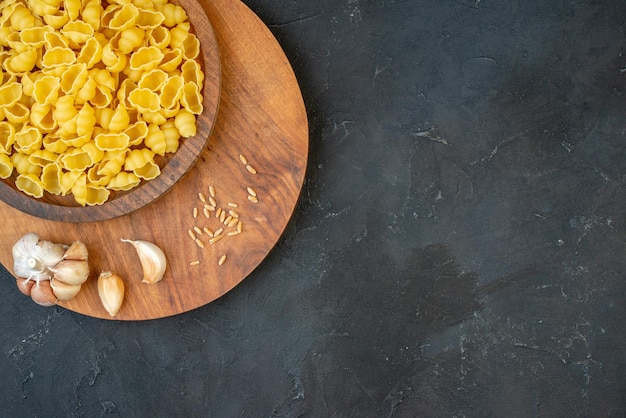 Overhead view of raw pastas in a brown bowl garlics rice on wooden board on the right side on black background