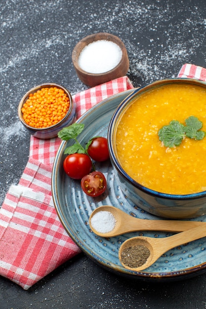 Overhead view of red lentil soup in a bowl served with greens tomatoes pepper salt on blue tray on red stripped towel and different spices on black white background