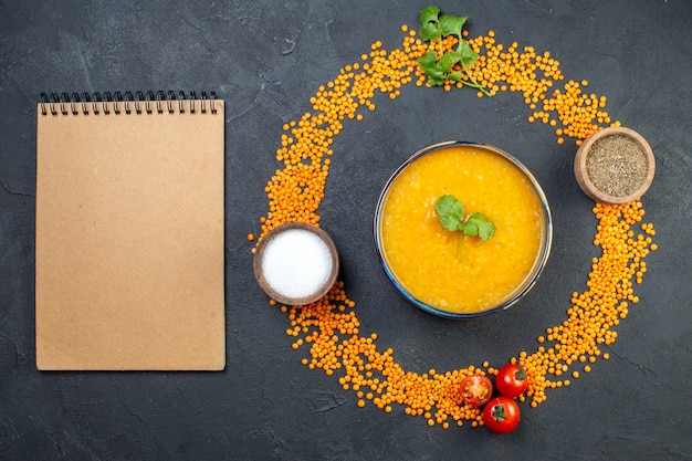 Overhead view of red lentils lined up in a circle and delicious soup salt pepper notebook on black background