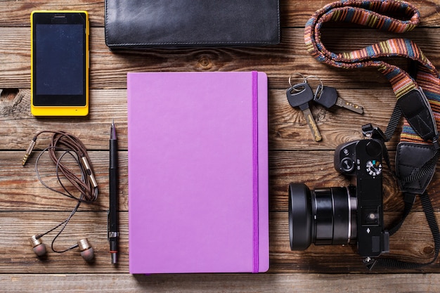 Overhead view of travel gear placed on wooden table. Mobile phone, earplugs, violet sketchbook, pencil, camera and purse. Flat lay top view.