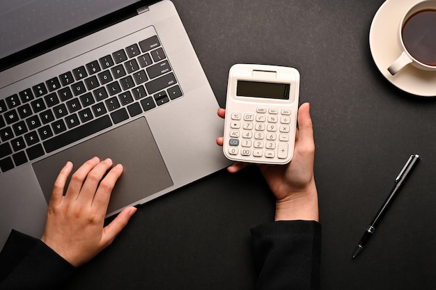 Photo overhead view workspace with a female holding a calculator and typing on a laptop computer on black table background