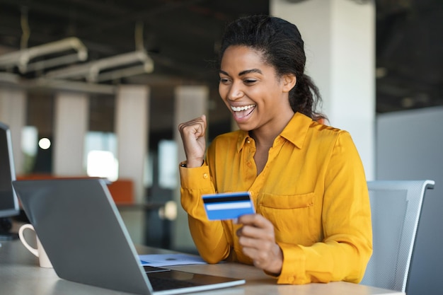 Overjoyed african american businesswoman making success gesture holding credit card sitting at table with laptop