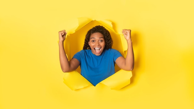 Overjoyed black woman cheering with clenched fists celebrating victory posing in torn hole of yellow background