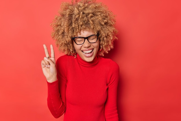 Photo overjoyed curly haired woman laughs happily makes peace gesture enjoys life keeps eyes closed being in good mood wears spectacles and turtleneck isolated over red background body language concept