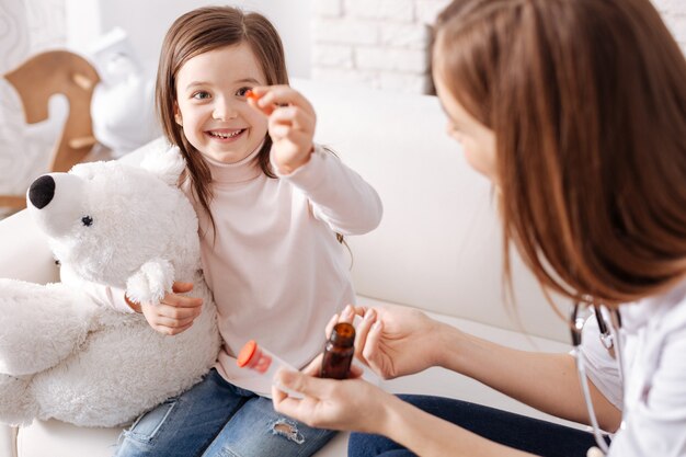 Overjoyed delighted little girl showing pill and going to take it while sitting on the couch with professional doctor