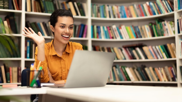 Overjoyed female teacher having online class using laptop and waving hand at webcamera panorama with free space