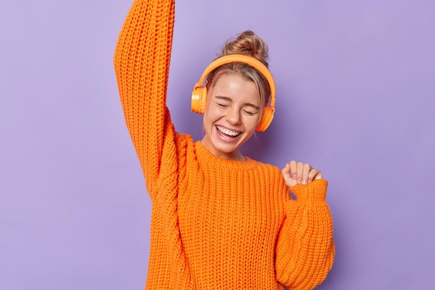 Overjoyed millennial girl shakes arms dances and has fun listens music in wireless stereo headphones wears warm knitted sweater isolated over purple background. People hobby happiness concept