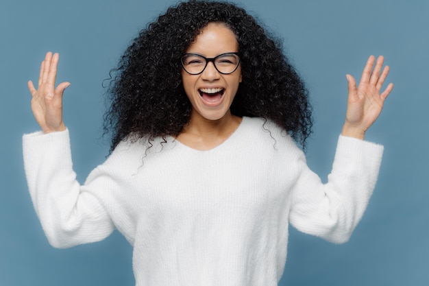 Overjoyed mixed race woman with curly hair raises hands exclaims from positive emotions keeps mouth opened dressed in white sweater stands against blue background Facial expressions concept