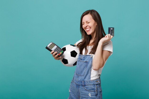 Overjoyed pretty young woman football fan support favorite team with soccer ball, wireless modern bank payment terminal to process, acquire credit card payments isolated on blue turquoise background.