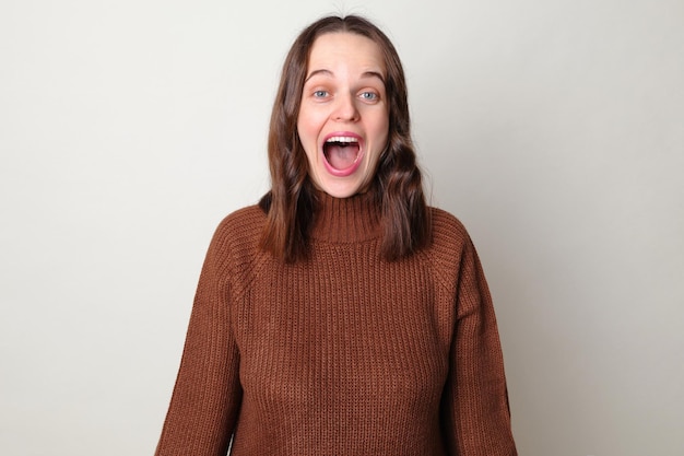 Overjoyed surprised brown haired adult woman wearing brown sweater posing isolated over gray background screaming with excitement and amazement with widely open mouth