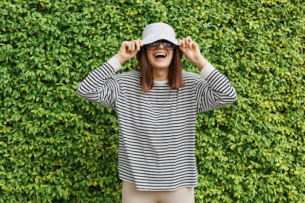 Overjoyed woman with brown hair wearing striped shirt panama and sunglasses posing near green garden fence looking at camera with excited amazed expression