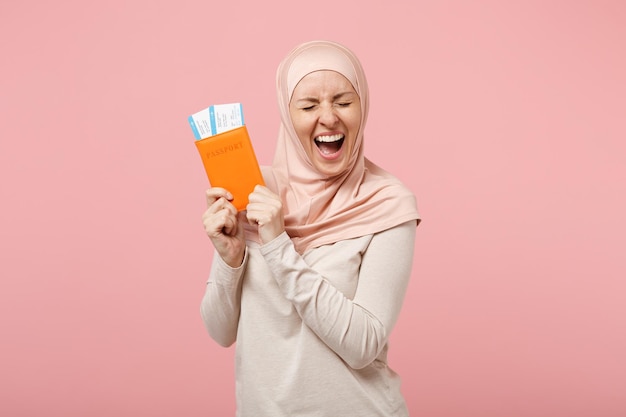 Overjoyed young arabian muslim woman in hijab light clothes posing isolated on pink background. People religious Islam lifestyle concept. Mock up copy space. Holding passport, boarding pass ticket.