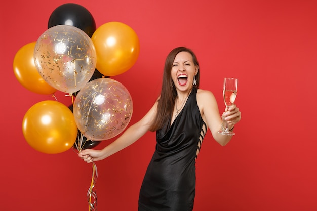 Overjoyed young girl in little black dress celebrating hold glass of champagne air balloons isolated on red background. International Women's Day Happy New Year birthday mockup holiday party concept.