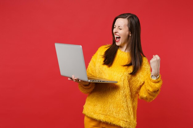 Overjoyed young woman in yellow fur sweater with closed eyes screaming, holding laptop pc computer isolated on red background in studio. People sincere emotions, lifestyle concept. Mock up copy space.