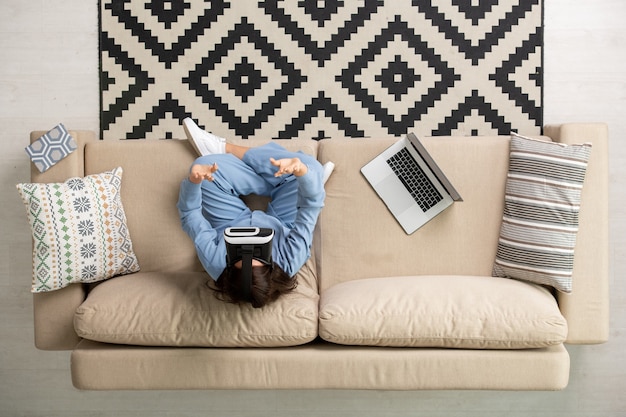 Overview of young brunette woman in blue pajamas and vr headset sitting on soft couch with her legs crossed and touching virtual display