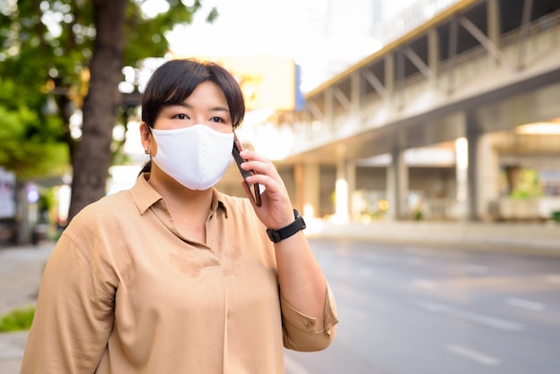 Overweight Asian woman with mask for protection from corona virus outbreak talking on the phone outdoors