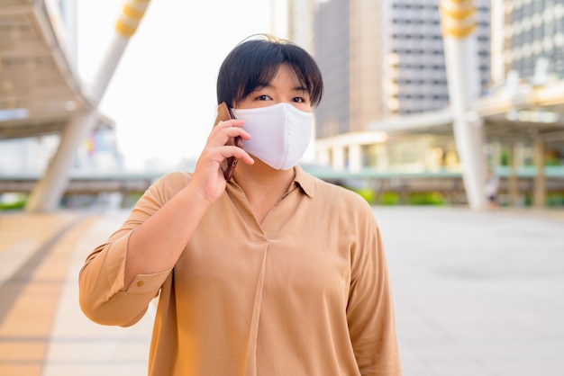 Overweight Asian woman with mask talking on the phone at skywalk bridge