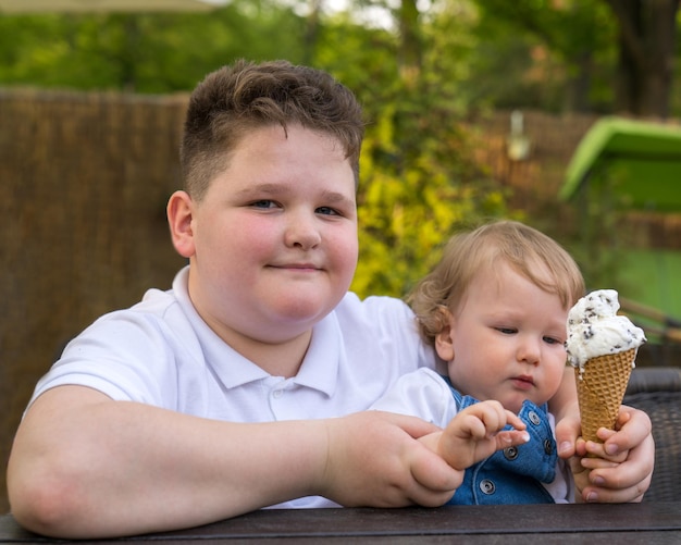 an overweight boy holds a fairhaired little child in his arms the kid eats ice cream