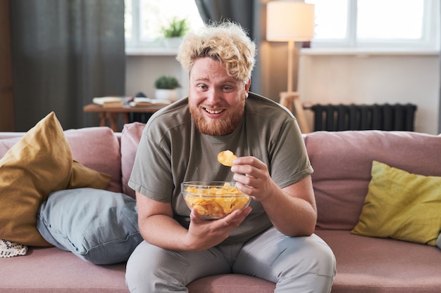 Overweight man sitting on sofa with bowl of chips eating them and laughing while watching tv at home