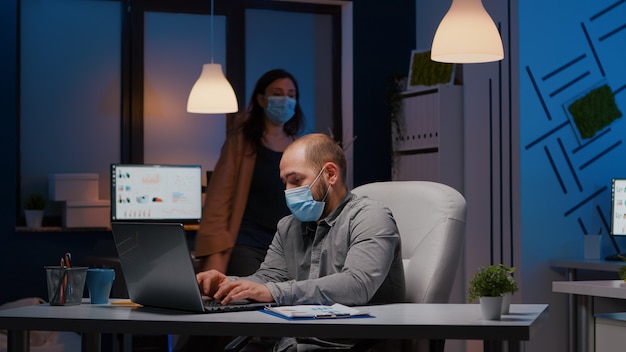 Overworked businessman with face mask against covid19 working in startup office checking management strategy late at night. Exhausted manager remains alone in company room after his collegue left