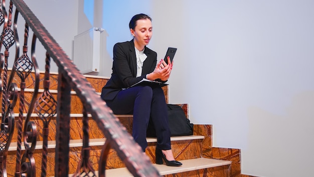 Overworked businesswoman smiling during video call about project deadline sitting on staircase of corporate company building. Entrepreneur talking with client using smartphone on stairs.