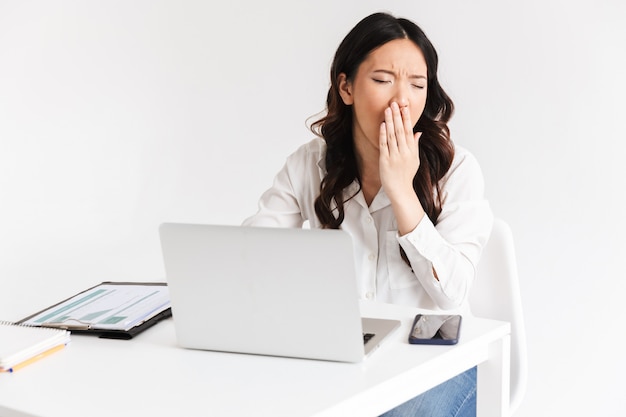 Overworked tired asian business woman wearing office clothing yawning while sitting at table with laptop open