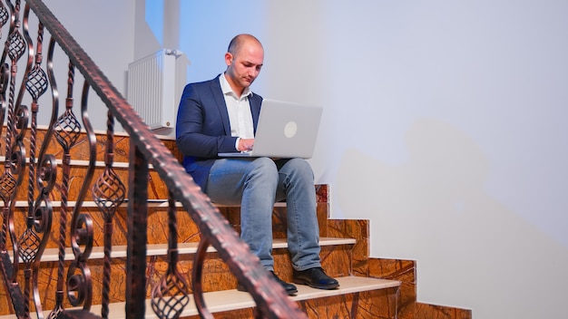 Overworked tired businessman at workplace working on deadline for job project using notebook sitting on stairwell. Confident corporate entrepreneur using laptop doing overtime in business building.