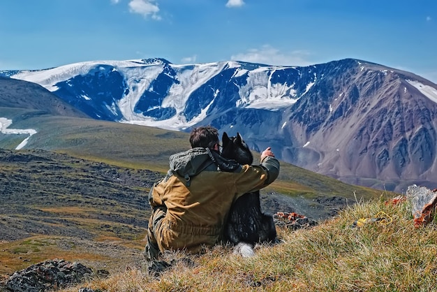 The owner with his beloved husky dog sits on a hill and points his hand to the mountains