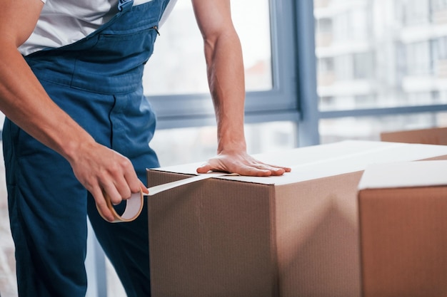 Packaging the box Young male mover in blue uniform works indoors in the room