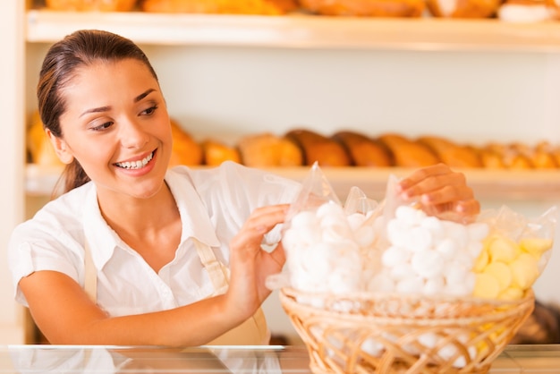 Packing fresh cookies for sale. Beautiful young woman in apron packing cookies and smiling while standing in bakery shop