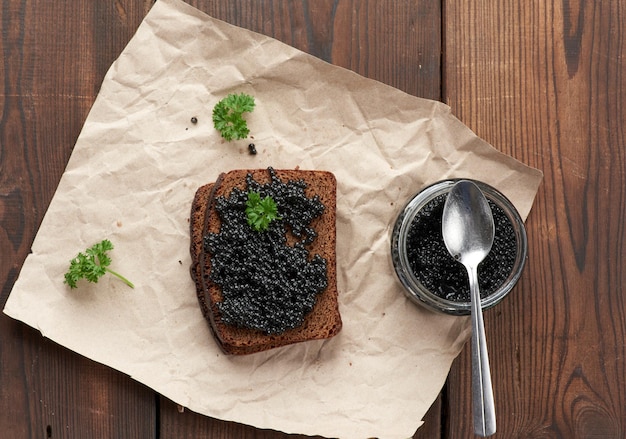 Paddlefish caviar, a slice of bread and a jar on a piece of brown paper, top view