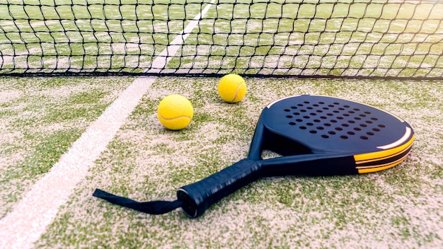 Padel racket and yellow ball behind net on a green court grass turf outdoors