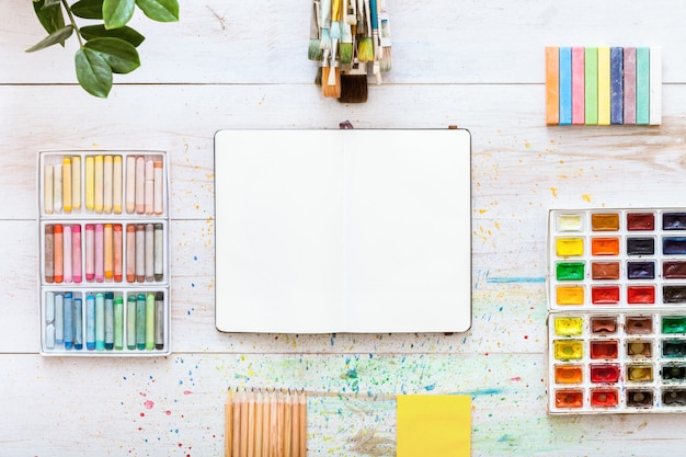 Paint brushes, paintbox watercolors, open notebook on white wooden background