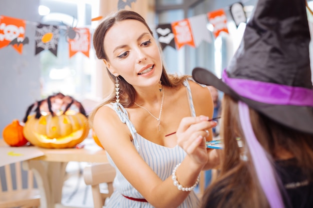 Paintbrush for face. Beautiful woman with nice makeup holding paintbrush while painting face of girl getting ready for Halloween