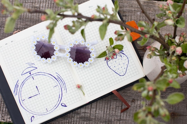 a painted alarm clock and a strawberry berry with a pen in a womens diary Summer country sketches in nature with an apple tree branch and sunglasses