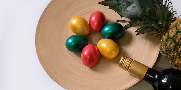 Painted easter eggs red green and gold colours with bottle red wine and pineapple on the white background, top view.