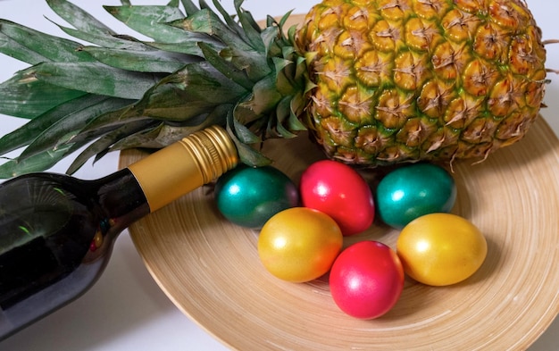 Painted easter eggs red green and gold colours with bottle red wine and pineapple on the white background, top view.